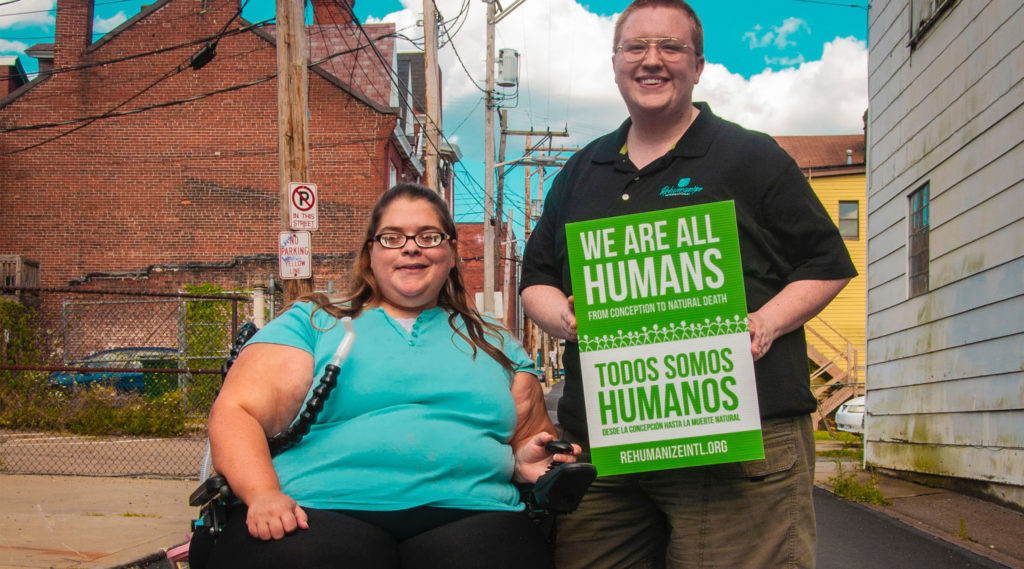 Two people smile, one standing and one in a wheelchair, and hold a sign that says "we are all humans from conception to natural death" in English and Spanish.