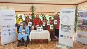 Family Safety Committee members smiling for a photo in a room with programme booklets, surrounded by pull-up banners showing the UN Spotlight Initiative logo