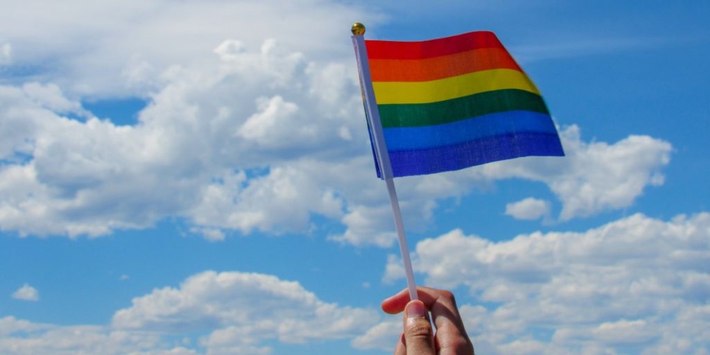 A hand holds a rainbow LGBT flag up to a cloudy blue sky