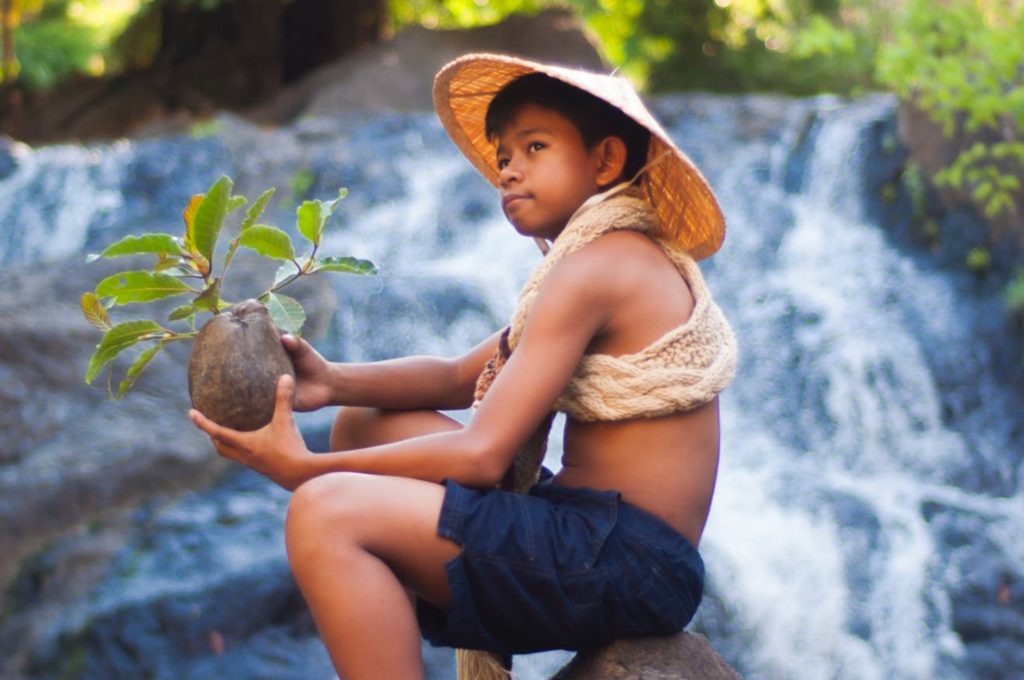 Young Asian boy sitting by a waterfall holding a sprouting coconut